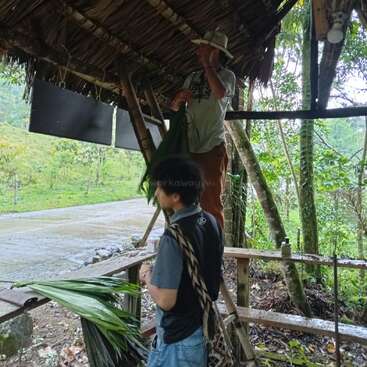 Two people under a rustic shelter with a thatched roof work with large green palm leaves. Lush greenery surrounds them, and they appear focused on their tasks.