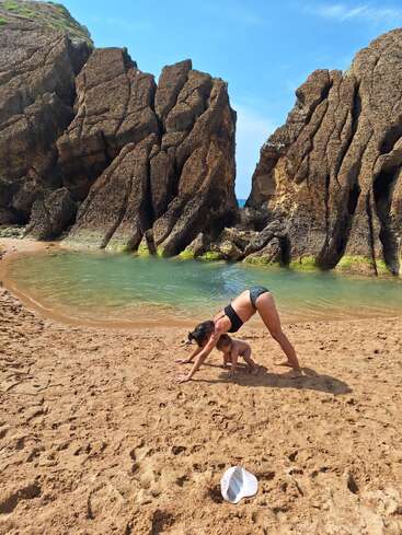 Una mujer y un niño hacen yoga en una playa de arena, con grandes rocas y una masa de agua al fondo, bajo un cielo azul.