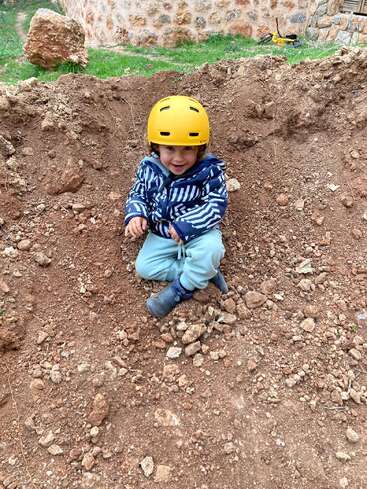 Un niño pequeño sentado en un agujero de tierra, con casco amarillo, chaqueta a rayas azules y blancas, pantalones azul claro y botas azules.