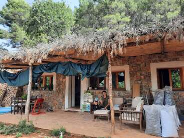 La imagen representa una serena casa de piedra con un porche cubierto, en la que aparecen una mujer y un niño, rodeados de frondosos árboles y un cielo azul, que desprenden una sensación de tranquilidad y calidez.