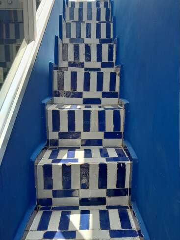 Una vibrante escalera con azulejos rectangulares azules y blancos en un patrón geométrico, flanqueada por paredes azules macizas e iluminada por la luz natural de una ventana.