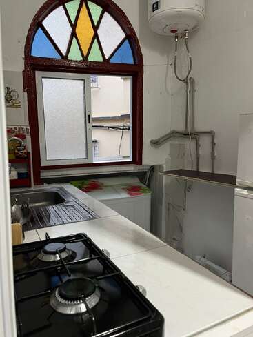 This small kitchen features a gas stove, sink, and washing machine. There's a unique arched stained glass window, white walls, and a wall-mounted water heater.