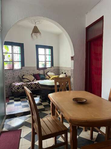 This cozy room has a wooden dining table with chairs, a checkered floor, arched entryway, cushioned daybed, patterned tiles, and sunlight streaming through window panes.