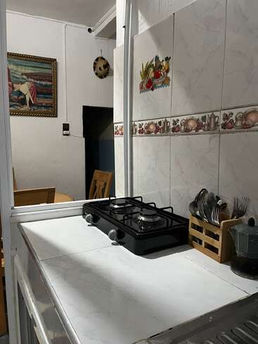 A small kitchen with a two-burner stove, utensil holder, Moka pot, white tiled countertop and walls. Background shows a dining table and painting.