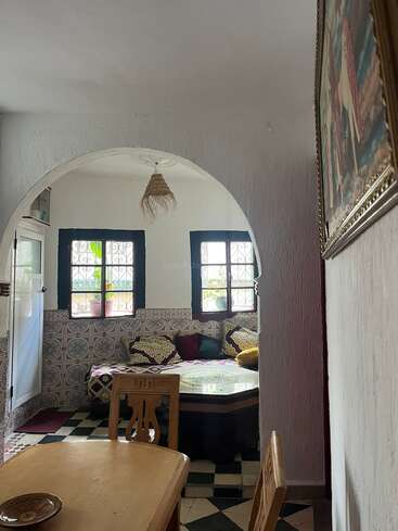 A cozy room with patterned tiles, arched doorway, wooden furniture, and a small sofa with colorful cushions. Two windows let in natural light. Warm, inviting atmosphere.