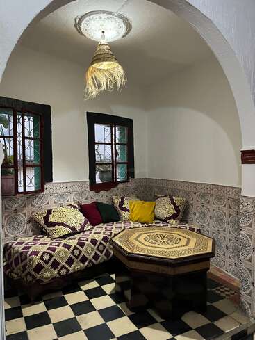 This cozy room features traditional Moroccan decor, patterned cushions, a geometric octagonal table, checkered floor tiles, detailed wall tiles, two windows, and a woven lampshade.