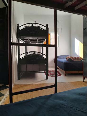 This image shows a simple, sunlit bedroom with metal bunk beds, a single bed, colorful pillows, a red rug, woven mat, and an open window.