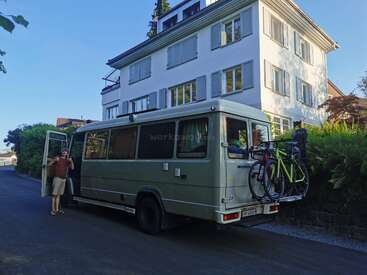 Ein Mann steht neben einem großen Wohnmobil, das in einer ruhigen Straße geparkt ist. Hinten sind zwei Fahrräder angehängt, im Hintergrund steht ein weißes Haus.