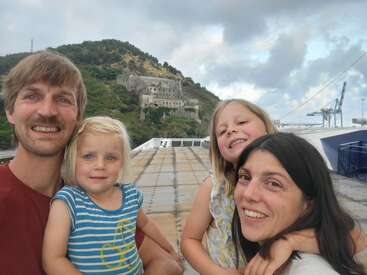 Eine lächelnde vierköpfige Familie - Eltern und zwei Töchter - posiert für ein fröhliches Selfie auf einer Fähre. Hinter ihnen tauchen ein grüner Hügel und ein historisches Gebäude auf.
