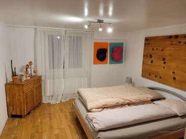 Dieses gemütliche Schlafzimmer verfügt über ein Doppelbett, zeitgenössische Kunst, Holzböden, eine Kommode mit Pflanzen, weiße Wände, Vorhänge und Deckenleuchten, die eine warme Atmosphäre schaffen.