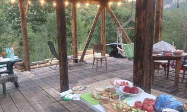 Une terrasse en bois chaleureuse, agrémentée de guirlandes lumineuses, de chaises disposées ici et là, d'un hamac, de deux tables garnies de plats, de tomates fraîches et de pain, le tout entouré d'arbres à la végétation luxuriante, en plein air.