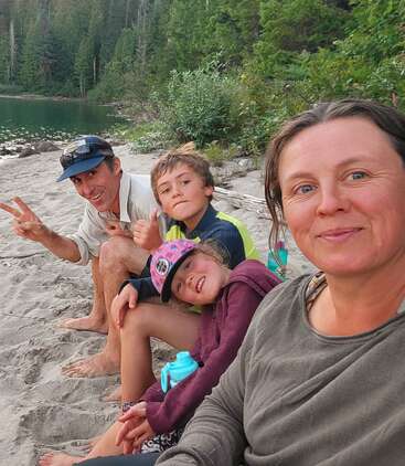 Eine vierköpfige Familie sitzt zusammen an einem Sandstrand am See, lächelt und genießt die Natur. Hohe grüne Bäume und das Wasser bilden eine friedliche Kulisse.