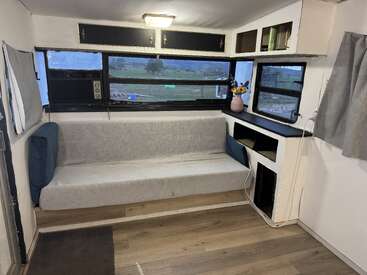 L'intérieur de ce camping-car confortable comprend une banquette coussinée grise, des armoires noires, des fenêtres avec vue sur la campagne, un plancher en bois, des étagères, un vase de fleurs et un éclairage doux.