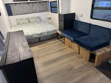 Cette image montre une chambre confortable et minimaliste avec un lit double, un canapé en bois à coussin bleu, une commode noire, des planchers en bois et plusieurs fenêtres avec des rideaux gris.