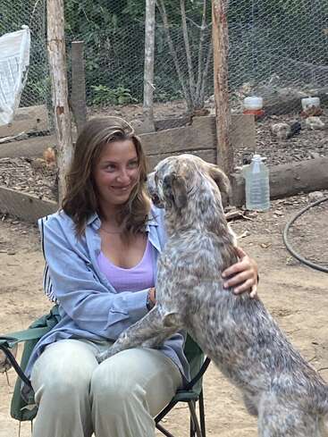 A young woman sitting on a chair outdoors smiles affectionately at a speckled dog standing on her lap. The atmosphere feels warm, rustic, and natural.