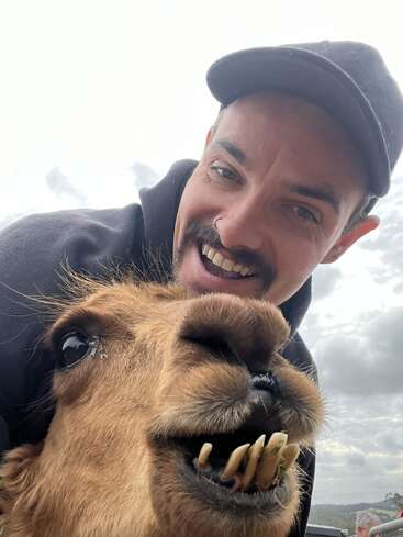 Un hombre con gorra sonríe alegremente junto a una llama que muestra sus grandes dientes. El fondo está nublado, creando un momento selfie juguetón y humorístico.