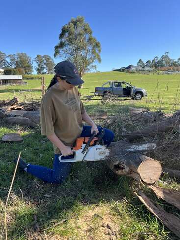 Una persona arrodillada sobre la hierba utiliza una motosierra Stihl para cortar un tronco grande. Hay un camión y un paisaje rural, que incluye árboles y una casa, al fondo.