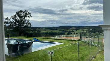 Una serena escena campestre con una piscina rodeada de vallas de cristal, exuberante hierba verde, colinas ondulantes, árboles lejanos, cielo nublado y apacible ambiente rural.