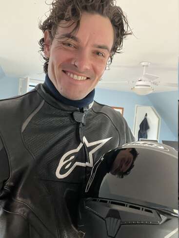 Uma pessoa sorridente vestindo uma jaqueta de couro preta de motociclista segura um capacete preto brilhante. Seu reflexo é visível no capacete. O fundo da sala inclui um ventilador de teto.