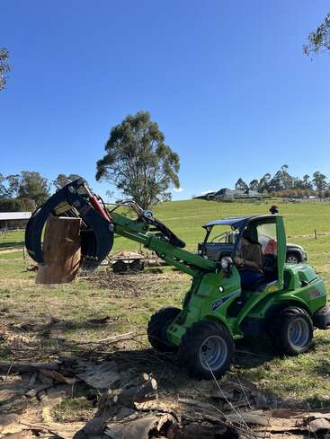 A person operates a small green loader with a claw attachment, lifting a large piece of wood on a sunny farm, surrounded by grassy fields and trees.