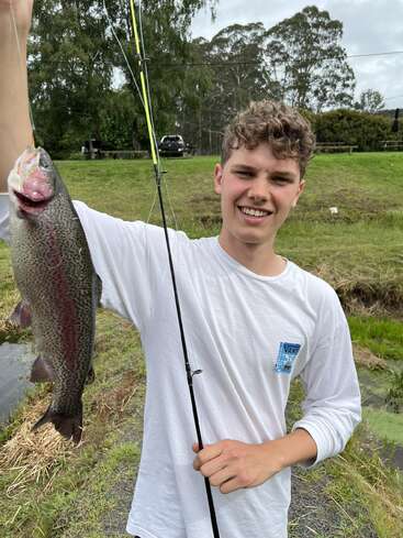 A smiling young man in a white shirt proudly holds up a large fish he caught, standing outdoors by a grassy, wooded area near a small stream.