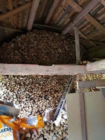 Das Bild zeigt einen Holzschuppen, der mit gestapeltem Brennholz gefüllt ist, mit einer Leiter, einer Tüte Hundefutter und einem Holzbalken, alles in einer rustikalen Holzkonstruktion.