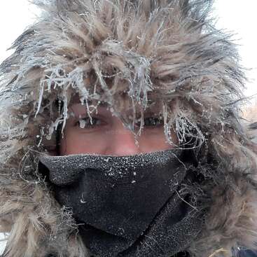 Das Bild zeigt das Gesicht einer Person, das teilweise von einem schwarzen Schal und einer pelzgefütterten Kapuze verdeckt wird, während Schneeflocken und Eiskristalle ihr Haar und ihre Kleidung bedecken.