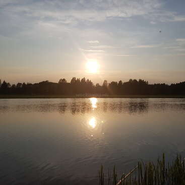 Das Bild zeigt eine ruhige Seeszene bei Sonnenuntergang mit der Spiegelung der Sonne auf dem Wasser und Bäumen am Horizont vor einem blauen Himmel mit Wolken.