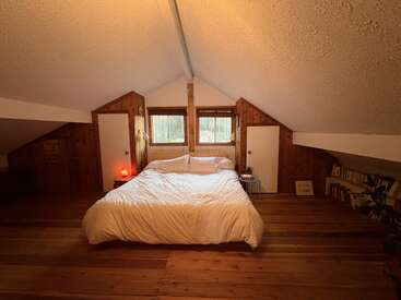 Cette chambre mansardée et chaleureuse est équipée d'un grand lit avec une literie blanche, de parquet et de murs en bois, d'un plafond en pente, de tables de chevet, de fenêtres, de livres et d'un éclairage d'ambiance chaleureux.