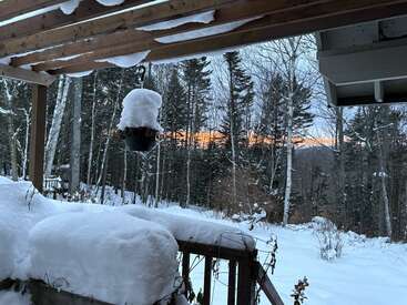 Une scène hivernale enneigée vue depuis une véranda couverte, avec des balustrades recouvertes de neige, des arbres en arrière-plan et un coucher de soleil orange vif qui pointe à travers la forêt. Une atmosphère paisible.