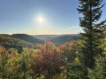 L'image montre un soleil éclatant au-dessus de collines vallonnées, une forêt dense au feuillage vert et rouge, ainsi qu'un ciel bleu limpide, évoquant une belle journée d'automne.