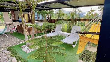 Eine gemütliche Outdoor-Lounge mit weißen Stühlen, einer gelben Hängematte, üppigen Grünpflanzen, einer überdachten Terrasse, einem Barbereich, Lichterketten und einer schönen Aussicht auf den Garten umgibt den Raum.