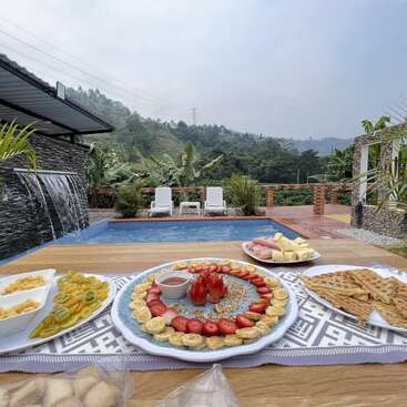 Ein köstliches Frühstück mit Obst, Waffeln und Käse wird auf einem Tisch am Pool serviert, umgeben von üppigem Grün und Bergen.