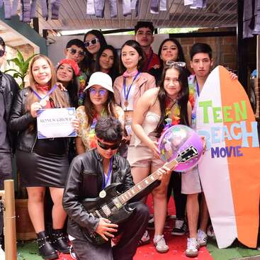 A group of teenagers dressed in beach and rock-themed attire pose with medals, a certificate, a guitar, and decorations inspired by "Teen Beach Movie."