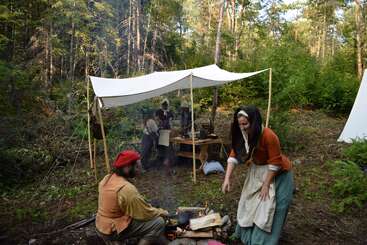 Varias personas vestidas con trajes de época acampan en un bosque. Dos de ellas avivan el fuego en primer plano, mientras que otras se reúnen bajo un toldo de lona, rodeadas de árboles y vegetación.