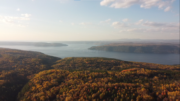Vista aérea de un paisaje con árboles teñidos de colores otoñales, colinas onduladas y un lago amplio y tranquilo bajo un cielo parcialmente nublado al atardecer. Una naturaleza preciosa.