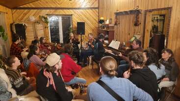 Un grupo de personas se reúne en una acogedora sala de madera y toca juntos diversos instrumentos musicales. El ambiente es cálido, acogedor y está lleno de alegría compartida.