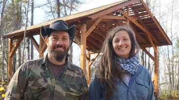 Un hombre con chaqueta y gorra de camuflaje y una mujer sonriente vestida de azul posan juntos al aire libre frente a una construcción de madera rodeada de árboles.