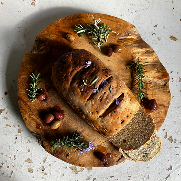 Das Bild zeigt ein rustikales Holzschneidebrett mit einem in Scheiben geschnittenen und mit Rosmarin und Oliven garnierten Laib Brot vor einem weißen Hintergrund.