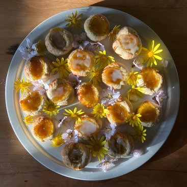 Das Bild zeigt einen weißen Teller mit einer Auswahl an kleinen Gebäckstücken, die jeweils mit einem Klecks Marmelade und essbaren Blumen auf einem Holztisch dekoriert sind.
