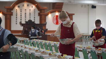 Várias pessoas usando aventais e máscaras estão preparando e servindo comida em muitos pratos dispostos em mesas compridas dentro de um salão grande e bem iluminado, com decoração tradicional.