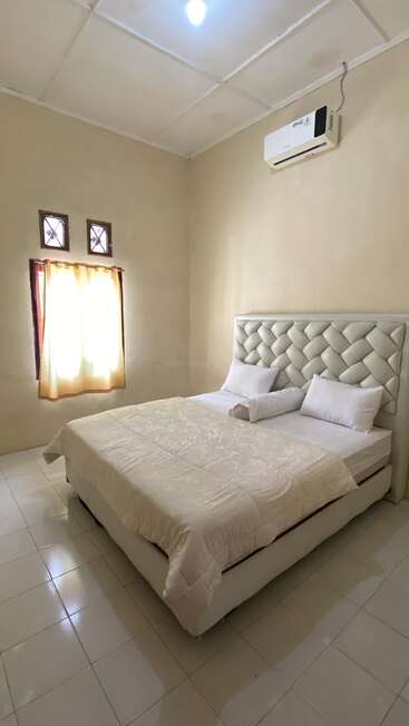 A simple, cozy bedroom with a large bed, tufted headboard, white bedding, small window with curtain, tiled floor, cream walls, and air conditioner. Minimalist decor.