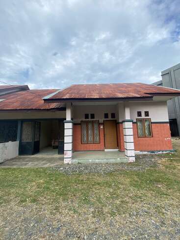 A single-story house with a rusty red roof, beige walls, and two pillars at the entrance. There is a small grassy yard and gravel in front.