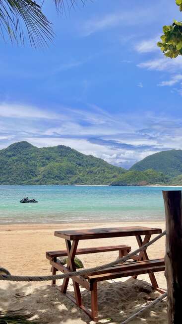A wooden picnic table sits on a sandy beach overlooking turquoise water, lush green hills, a lone jet ski, and a bright, partly cloudy sky above.