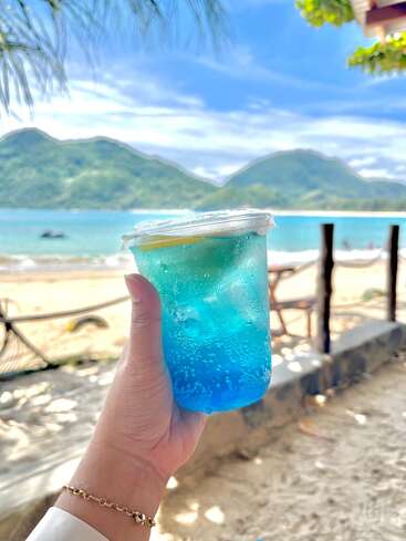 Une main tient une boisson bleue rafraîchissante avec de la glace et du citron, avec vue sur une belle plage, une mer turquoise et des montagnes vertes pittoresques sous un ciel ensoleillé.