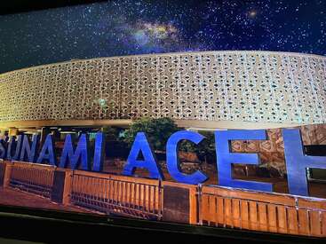 L'image montre le musée du tsunami d'Aceh de nuit, illuminé sous un ciel étoilé, avec de grandes lettres bleues épelant "TSUNAMI ACEH" devant le bâtiment.