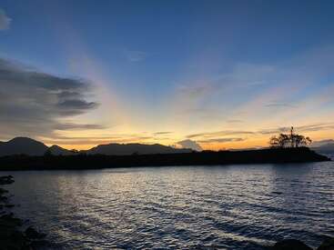 Une scène de coucher de soleil paisible sur des eaux calmes, avec des silhouettes de montagnes, une île avec des arbres, et un ciel paisible passant doucement du jaune d'or au bleu profond.