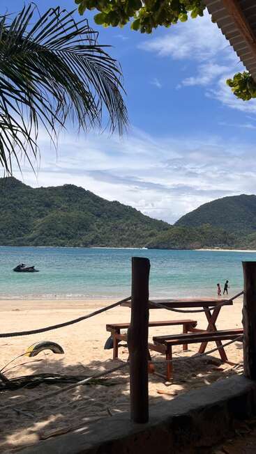 Une scène de plage paisible avec une table de pique-nique en bois, un rivage sablonneux, une mer bleue claire, des collines vertes, des palmiers, un jet-ski et deux personnes marchant près de l'eau.