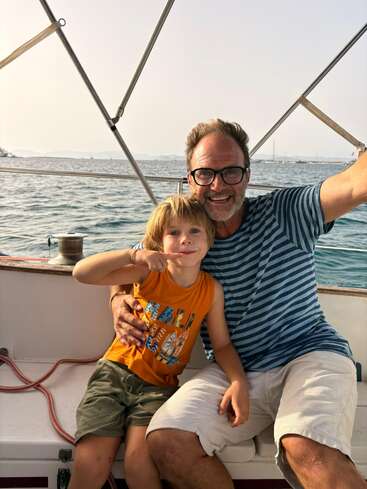 A man and a young boy are happily sitting together on a boat, smiling at the camera, with the ocean and distant boats in the background.