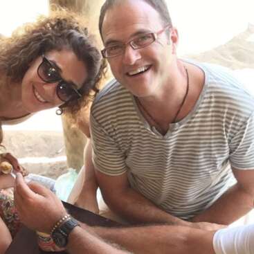 A man and woman smiling brightly at the camera. The man wears glasses and a striped shirt, the woman has curly hair and sunglasses. Casual, cheerful moment shared.